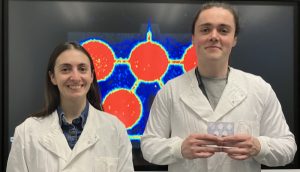 Dr Adriana Tavares and Liam Carr with the body on chip device standing in front of a PET image of the chip